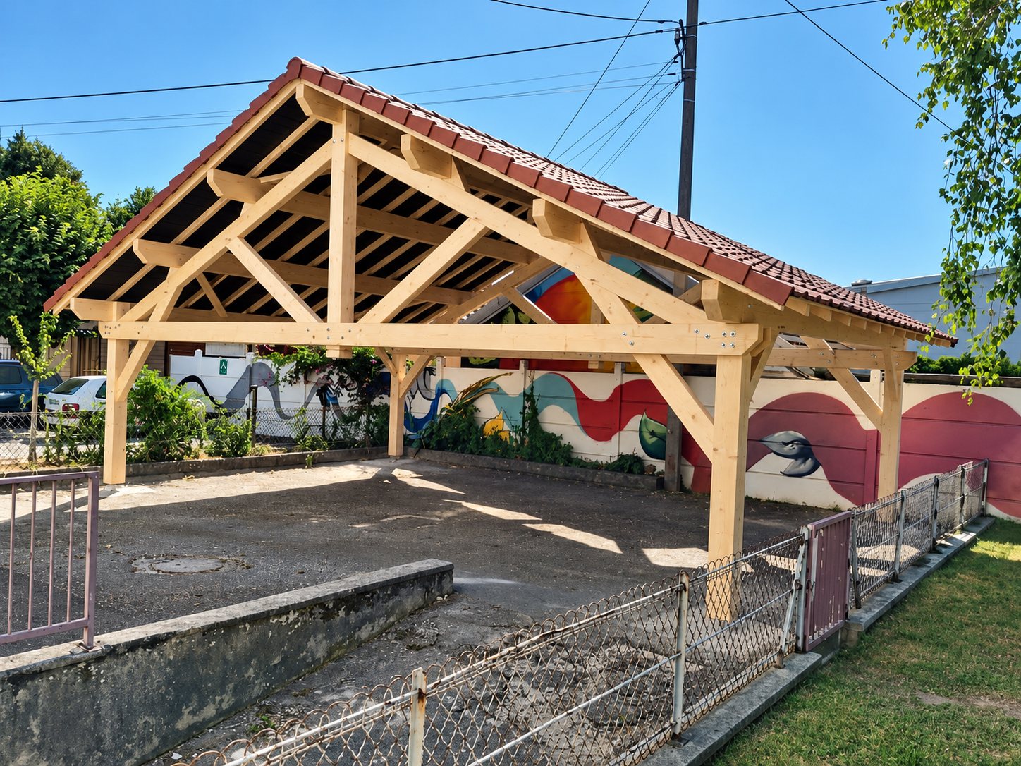 Un grand abri en charpente de bois clair couvert de tuiles rouges, installé sur une cour en terre devant un mur décoré d'une fresque colorée.