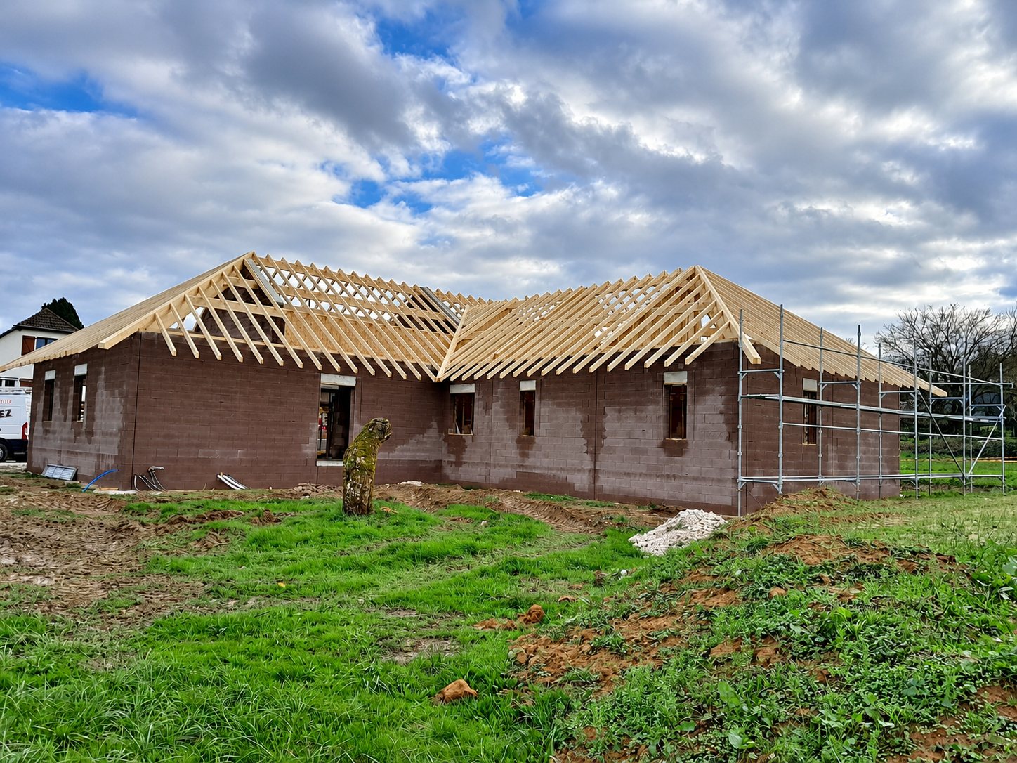 Chantier d'une maison de plain-pied en briques rouges dont la charpente en bois est posée, avec un échafaudage sur le côté droit et de l'herbe au premier plan.