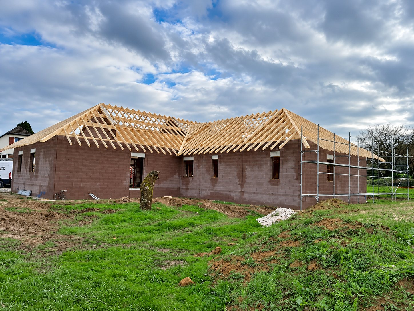 Une maison de plain-pied en cours de construction, avec ses murs en parpaings, sa charpente en bois posée sans toiture et un échafaudage sur le côté.