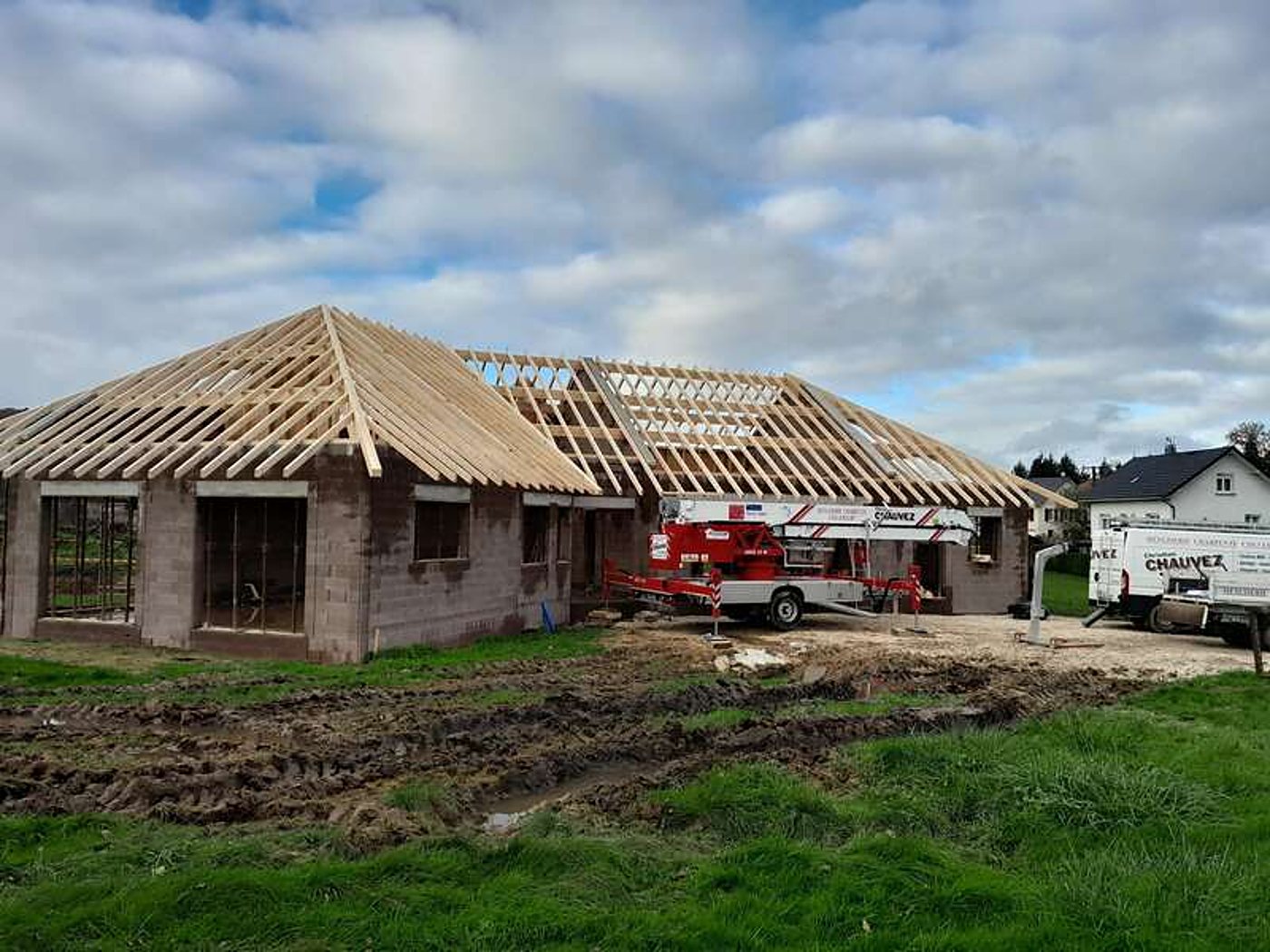 Chantier d'une maison en parpaings dont la charpente en bois est posée, avec une nacelle rouge et une camionnette de l'entreprise Chauvez à côté.