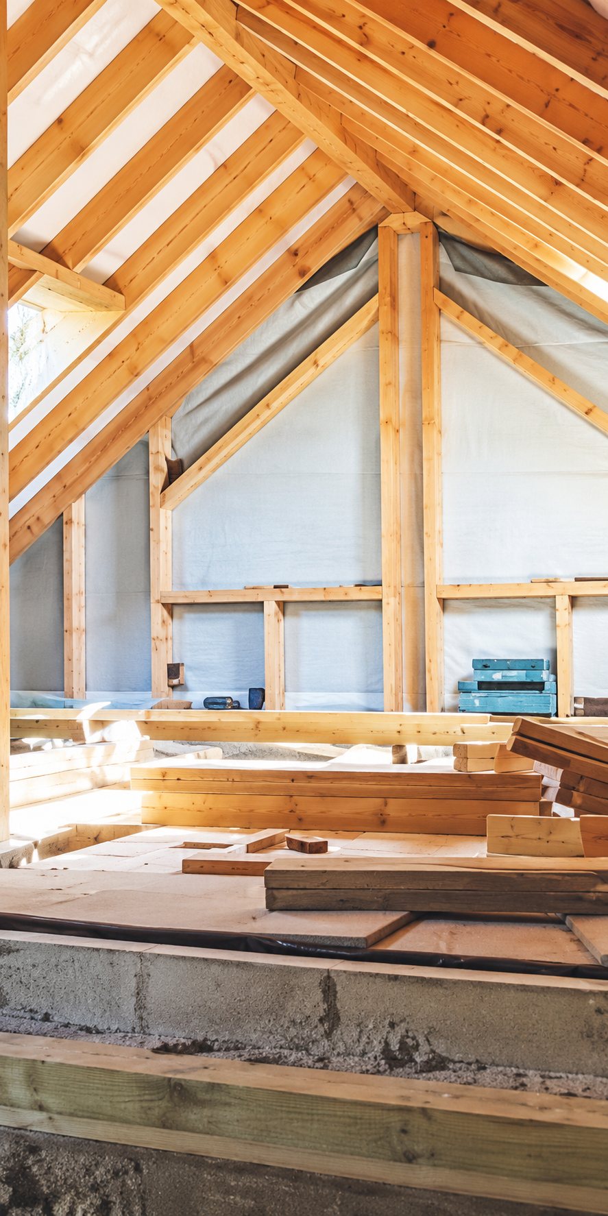 Vue intérieure d'un chantier de construction montrant une charpente en bois claire, des murs en cours d'isolation et des planches empilées au premier plan.