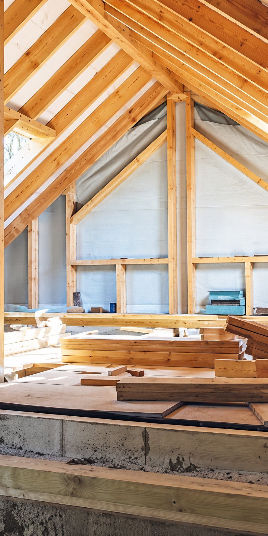 Intérieur d'une maison en chantier montrant la charpente en bois, les chevrons et des planches empilées au premier plan.