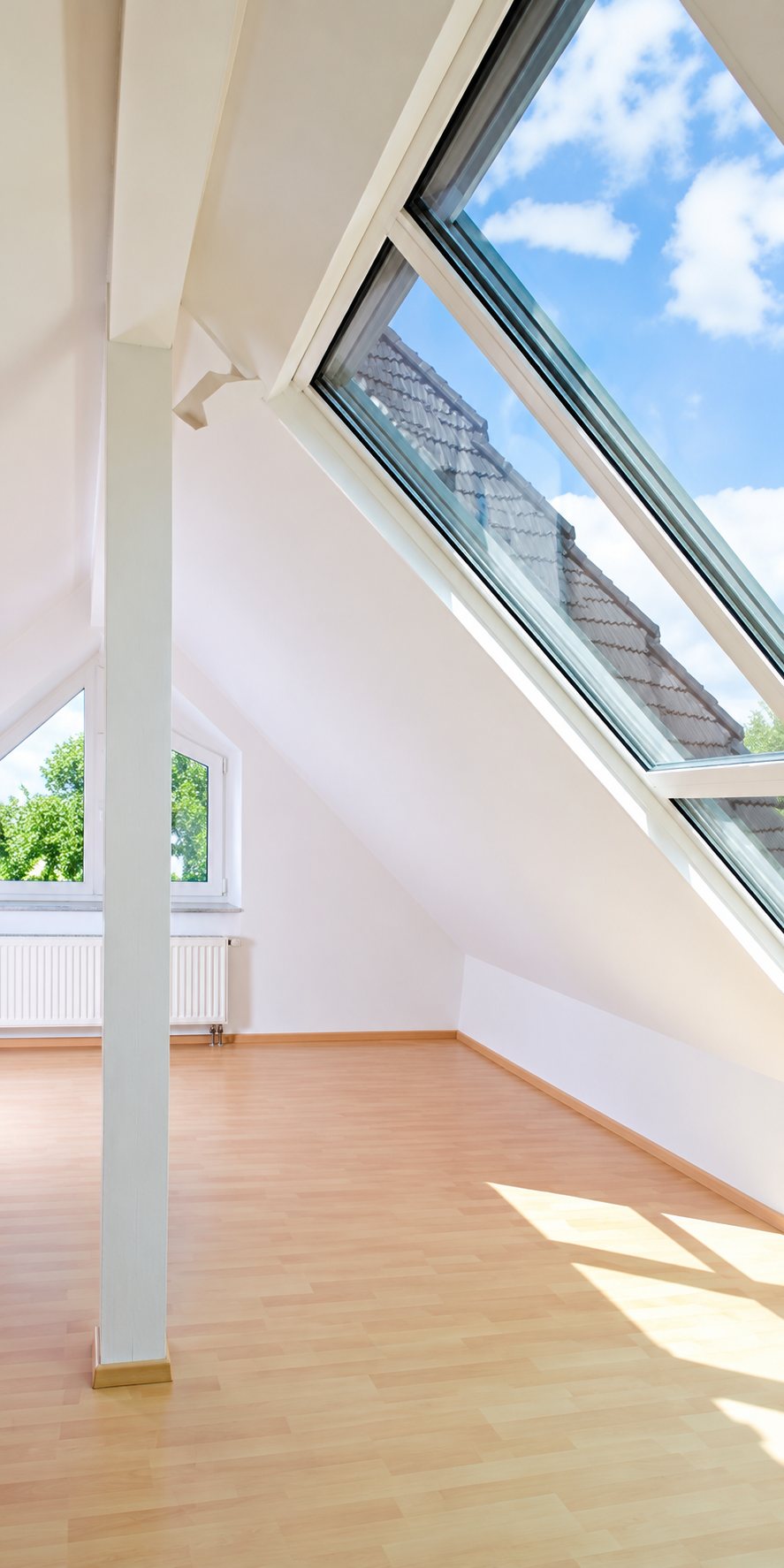 Pièce sous les toits rénovée avec grandes fenêtres de toit, poutre blanche et parquet en bois clair baigné de lumière.