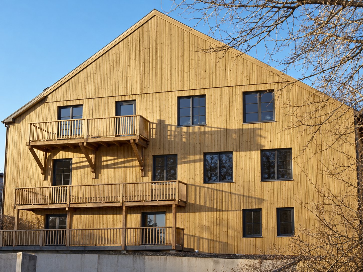 Façade d'un grand bâtiment revêtu de bardage en bois clair, avec fenêtres et balcons, éclairée par le soleil sous un ciel bleu.