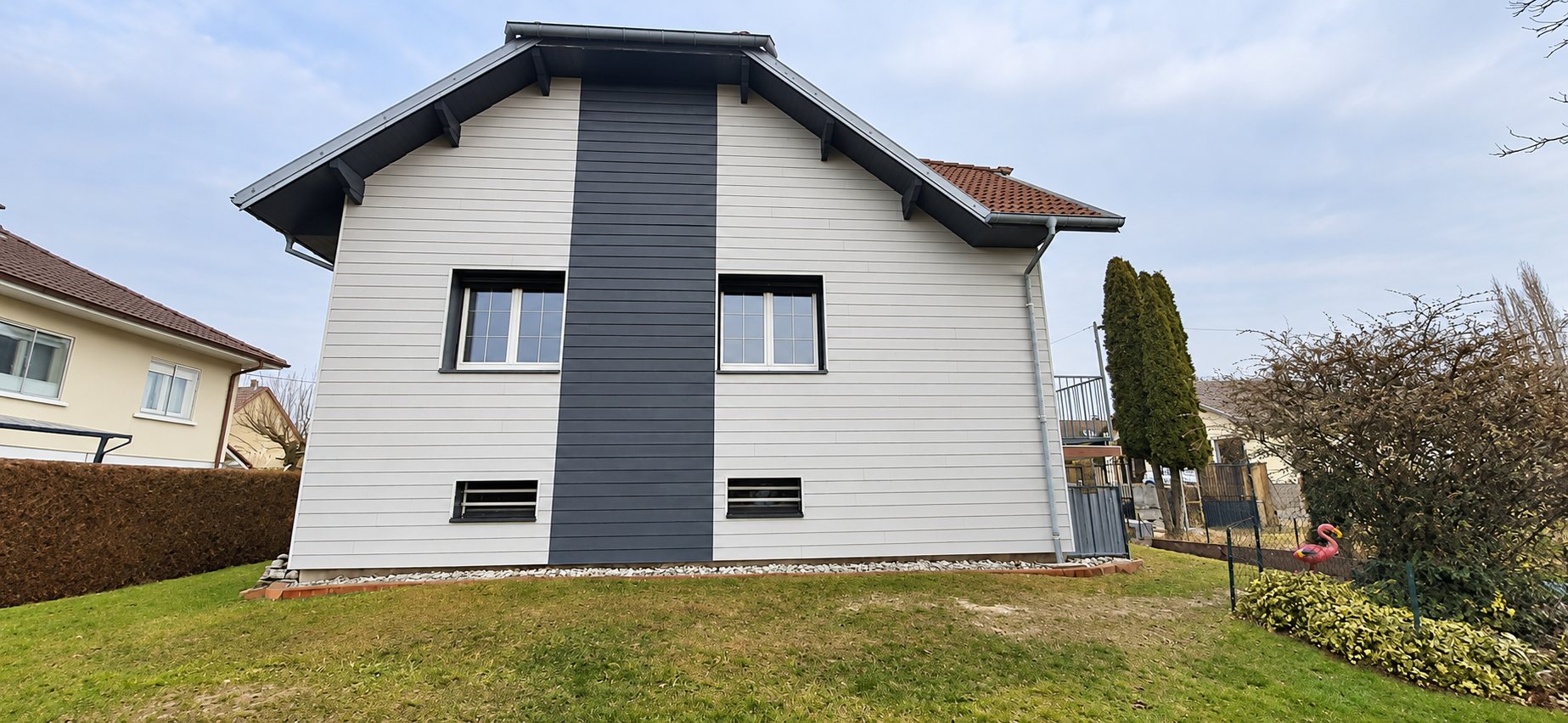 Façade d'une maison individuelle recouverte d'un bardage horizontal gris clair avec une large bande verticale gris anthracite entre deux fenêtres, sous un toit à pignon.