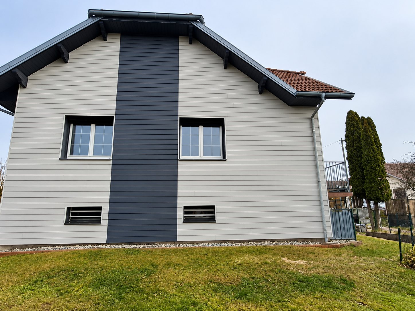 Façade d'une maison récemment rénovée avec un bardage horizontal gris clair, une large bande centrale anthracite, deux fenêtres blanches et un toit en tuiles.