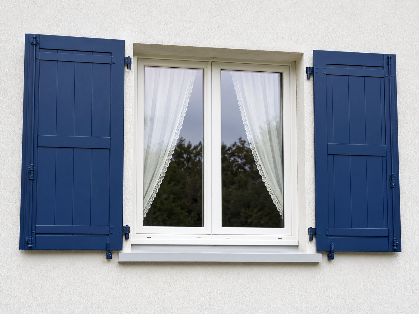 Une fenêtre blanche à deux battants avec des rideaux légers, encadrée par deux volets bleu foncé ouverts sur un mur clair.