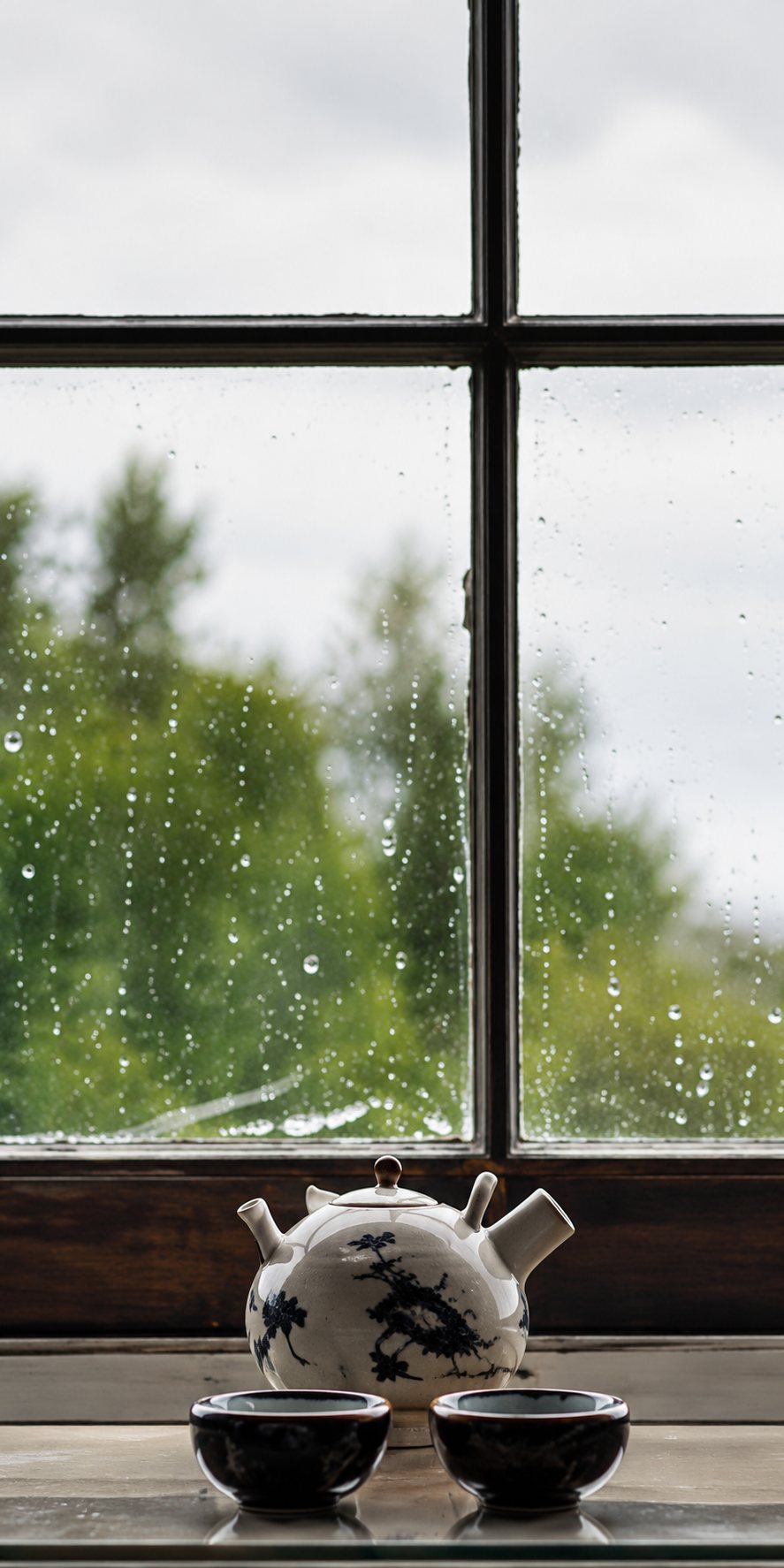Une petite figurine claire et deux bols sombres posés sur un rebord devant une grande fenêtre couverte de gouttes de pluie.