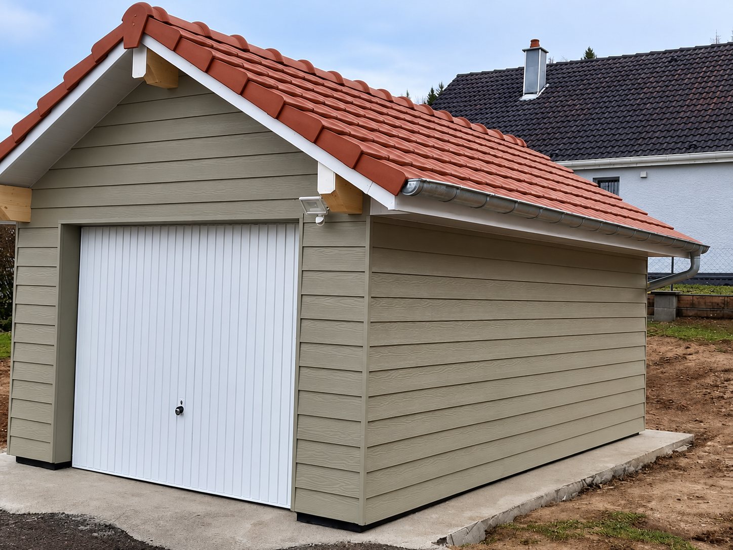 Garage récemment construit avec bardage horizontal beige, porte basculante blanche et toiture en tuiles rouges, posé sur une dalle en béton.