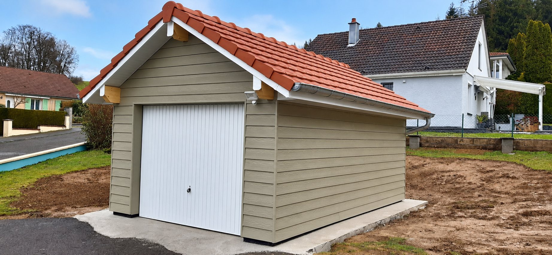Garage individuel récemment construit, avec bardage beige, porte basculante blanche et toiture en tuiles rouges, posé sur une dalle en béton dans un lotissement.