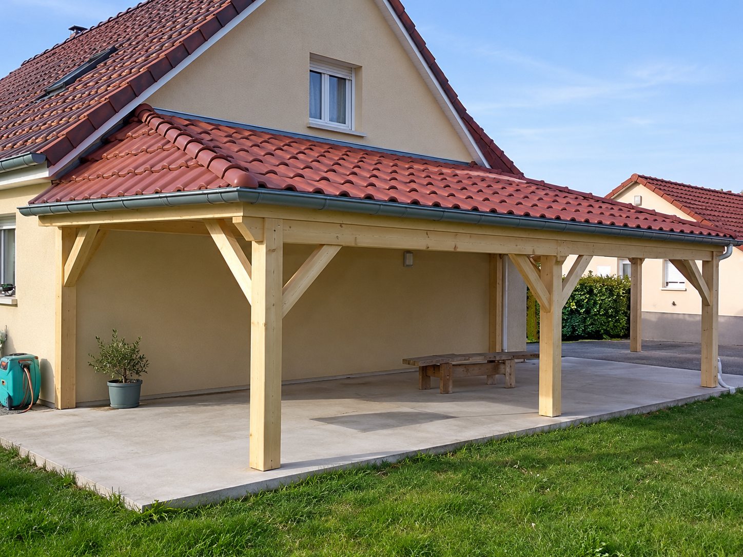 Une maison à toit de tuiles rouges prolongée par un auvent en bois couvrant une terrasse en béton, sous un ciel bleu.