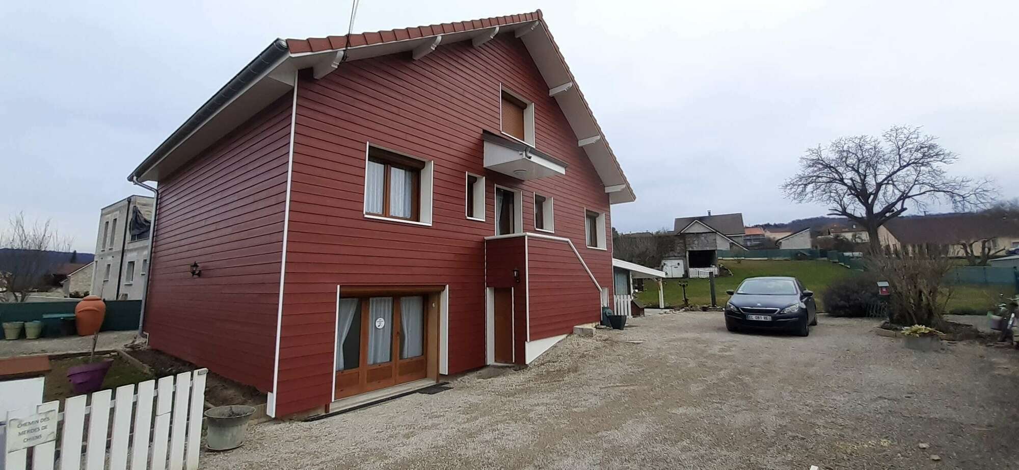 Maison à deux niveaux habillée d'un bardage rouge aux encadrements blancs, avec une voiture noire garée sur une allée de gravier.