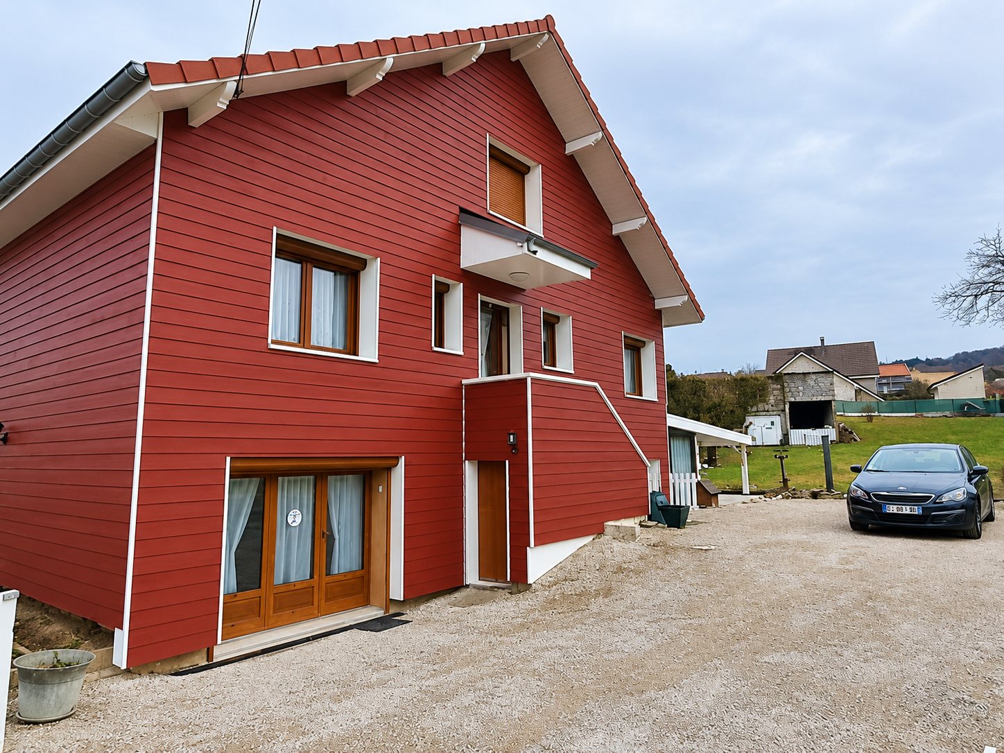 Maison à pignon dont la façade est recouverte d'un bardage rouge, avec fenêtres en bois et une voiture grise garée sur le gravier.
