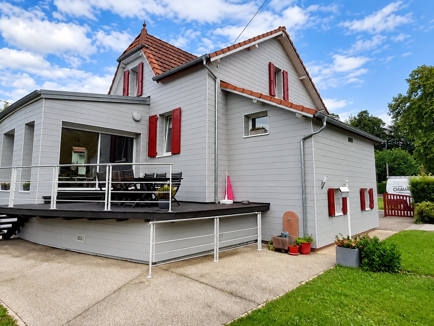 Maison à bardage gris clair avec volets rouges et une grande terrasse en bois surélevée, entourée d'une pelouse et d'un jardin.
