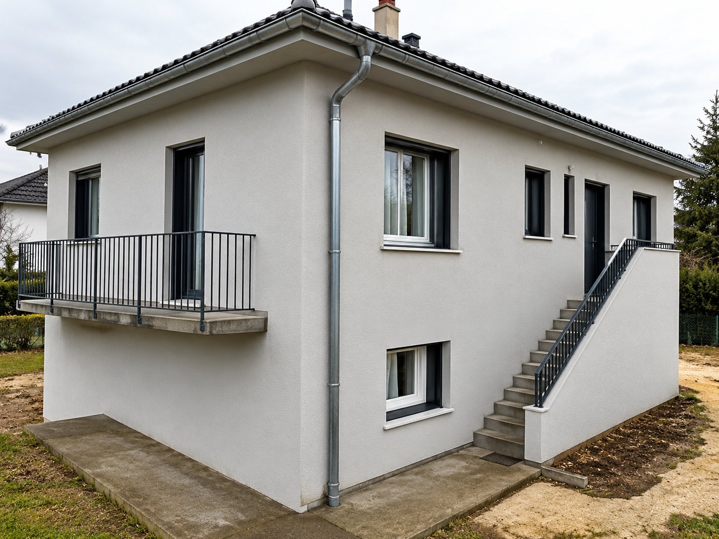 Une maison récente aux façades blanches avec un balcon à garde-corps métallique et un escalier extérieur, sous un ciel nuageux.