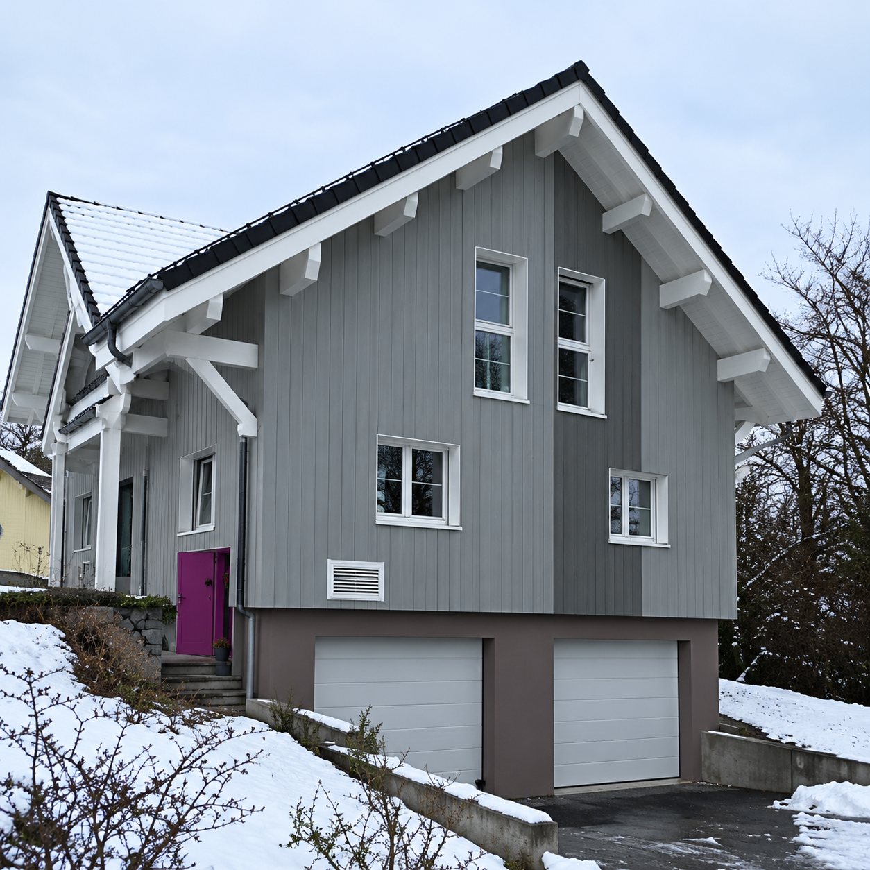 Une maison moderne aux façades grises et blanches, dotée d'un garage au rez-de-chaussée, se dresse sur un terrain en pente légèrement enneigé.