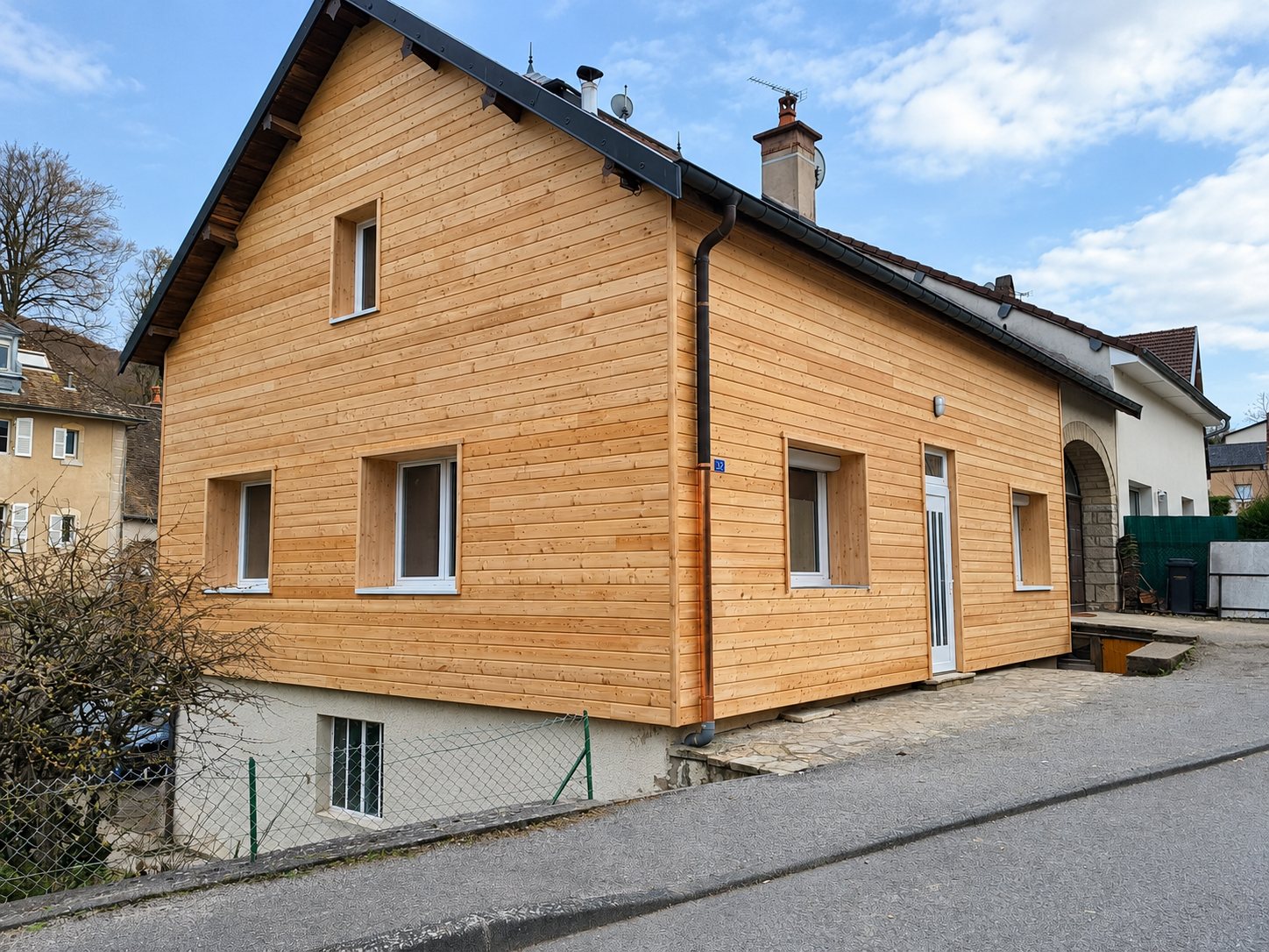 Une maison dont la façade a été rénovée avec un bardage horizontal en bois clair, vue depuis la rue, avec des fenêtres blanches et une porte blanche.