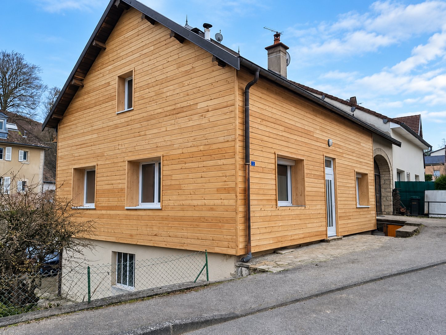 Maison individuelle récemment rénovée dont les façades sont recouvertes d'un bardage en bois clair horizontal, avec un toit gris foncé et des fenêtres blanches, au bord d'une rue.