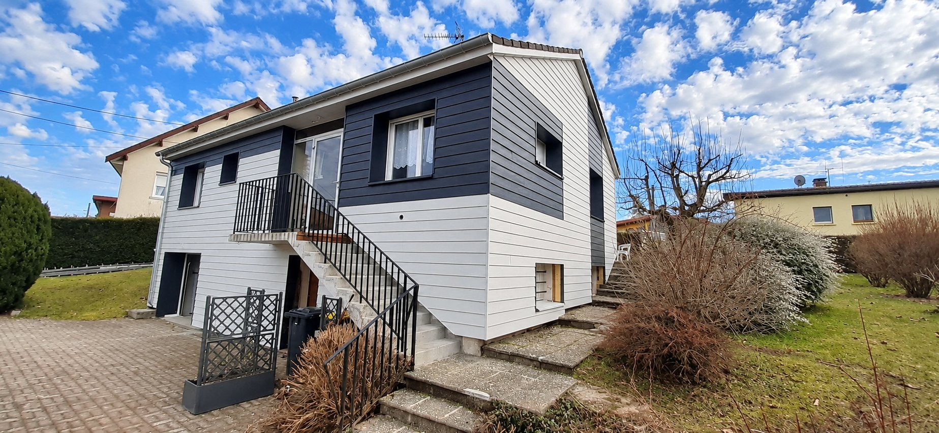 Maison individuelle récemment rénovée avec un bardage gris clair et anthracite, un escalier extérieur à rambarde noire et un jardin autour.
