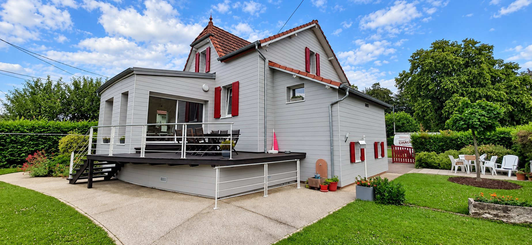 Maison rénovée au bardage gris clair avec volets rouges et grande terrasse en bois surélevée, entourée d'un jardin et d'une allée en gravillons.