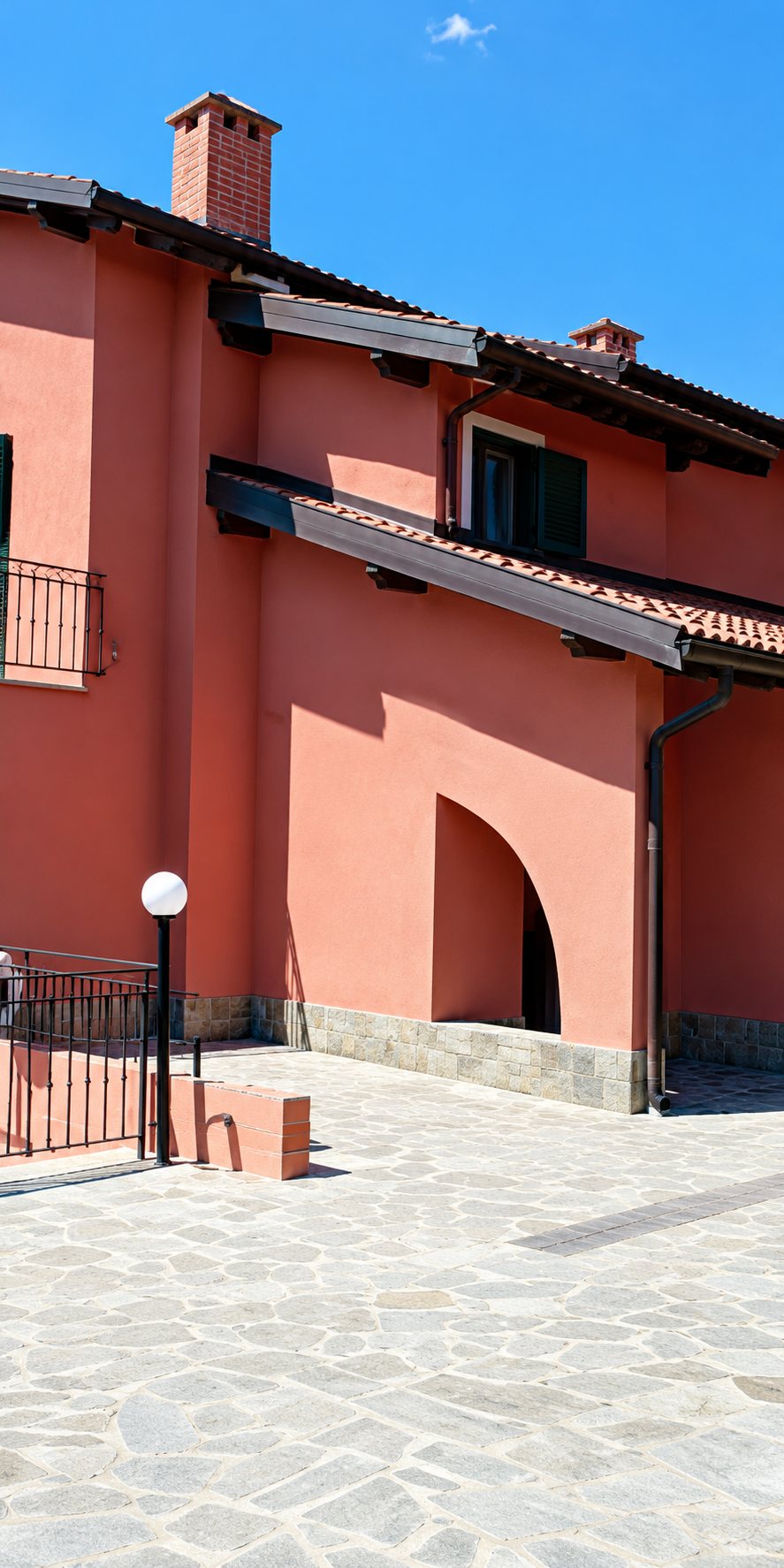 Façade d'un bâtiment rose à toit brun avec une arche et une cour pavée de pierres claires au premier plan.
