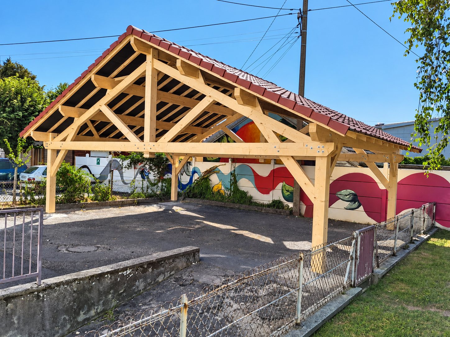 Un grand abri en bois neuf avec charpente apparente et couverture de tuiles rouges, installé devant un mur décoré d'une fresque colorée.