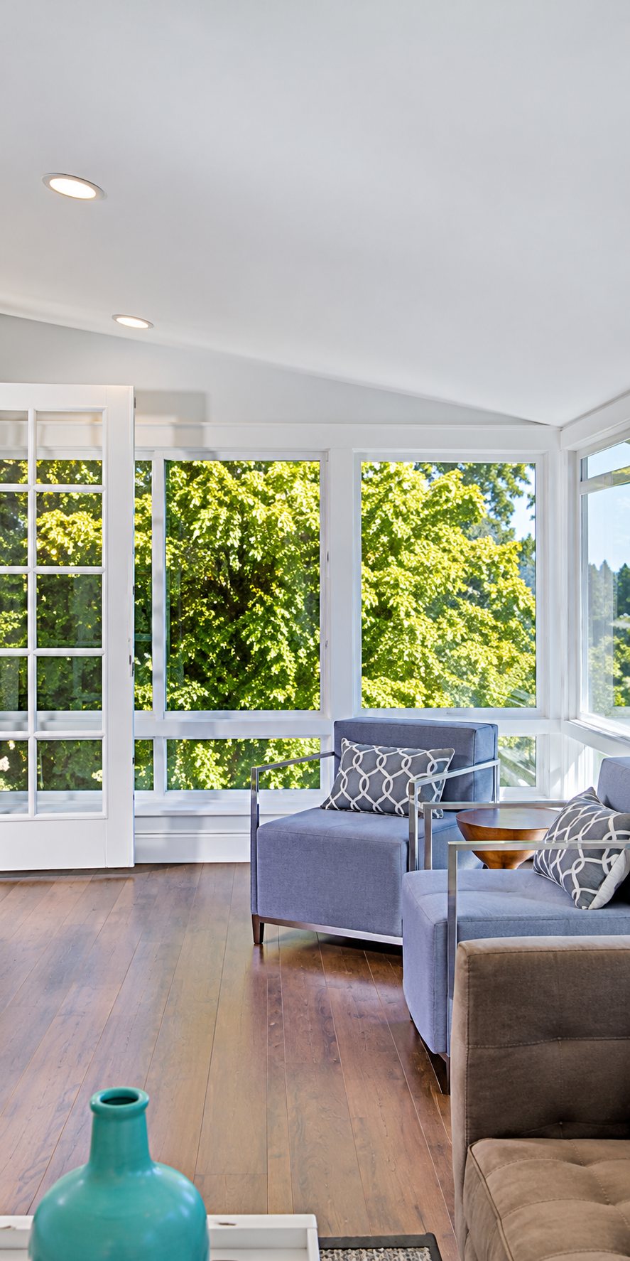 Un salon lumineux au parquet clair avec un fauteuil bleu lavande, un canapé gris et de grandes fenêtres donnant sur la verdure, avec un vase turquoise au premier plan.