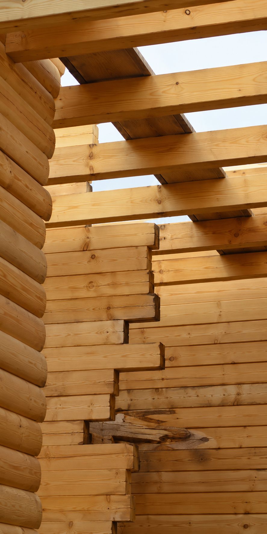 Angle d'une construction en rondins de bois empilés, avec des poutres en bois apparentes au-dessus et le ciel visible entre elles.
