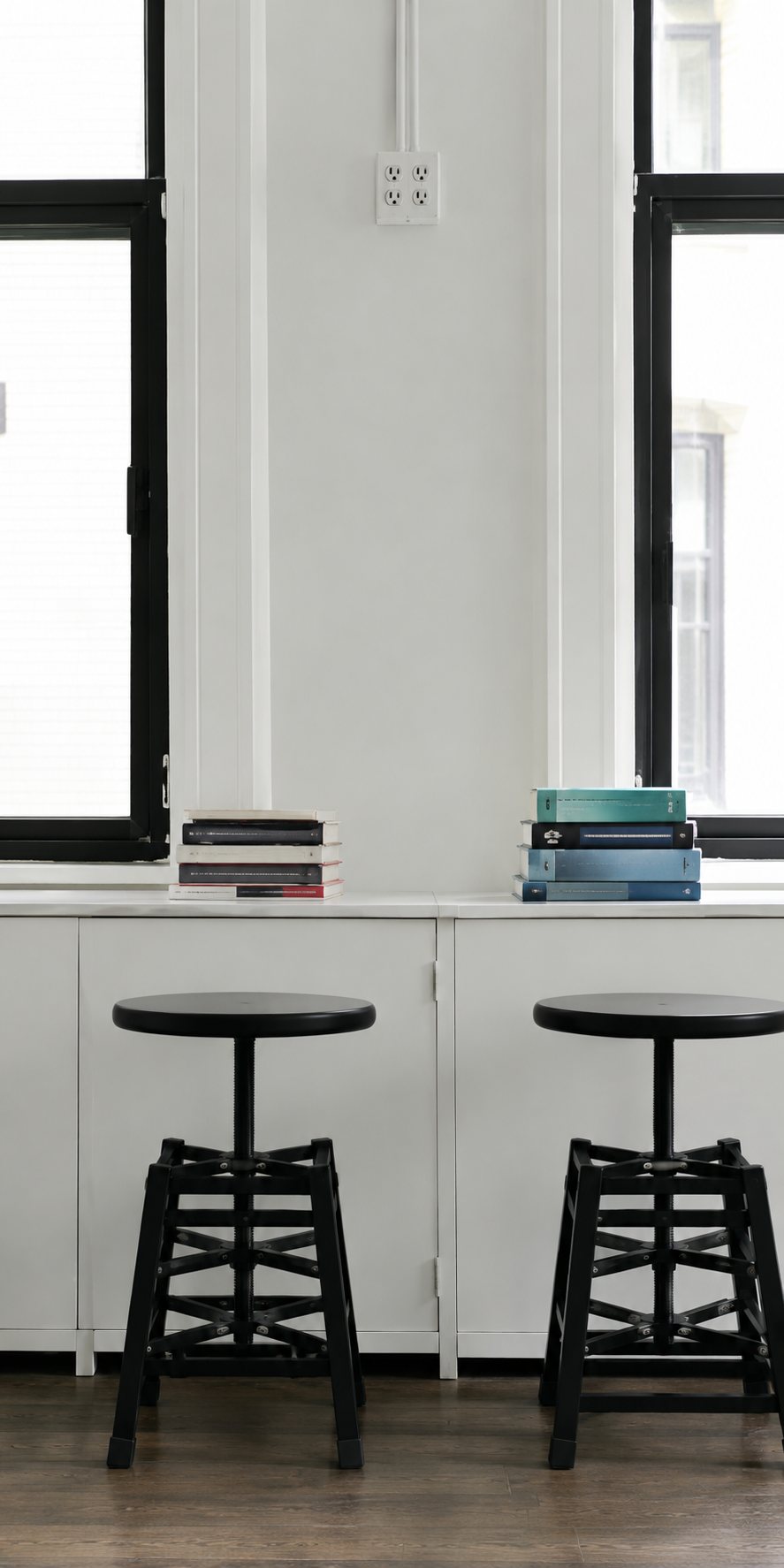 Deux tabourets en bois noir sont alignés devant un long comptoir blanc, sous de grandes fenêtres à cadre noir, avec des piles de livres posées dessus.